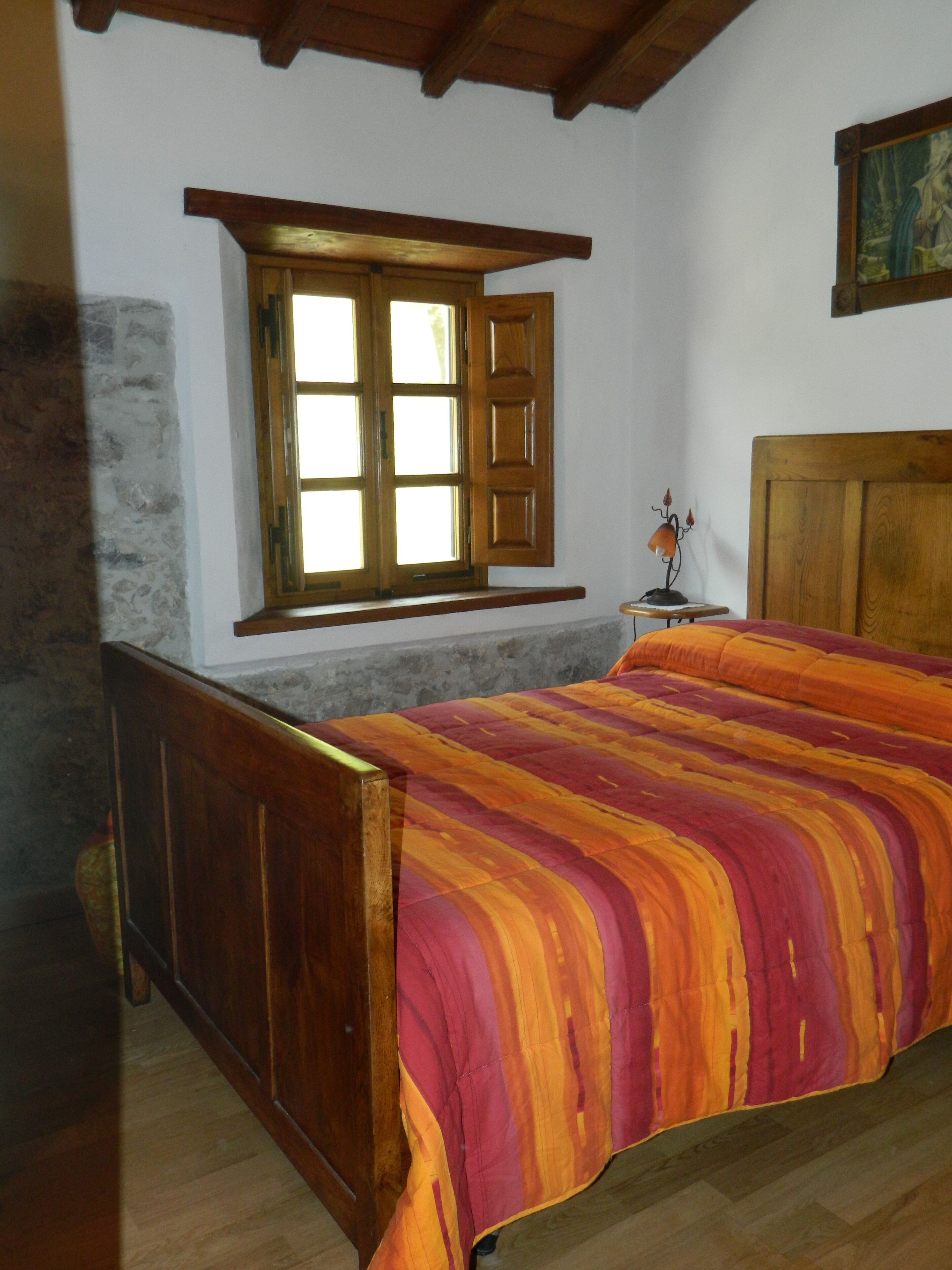 A bedroom with a wooden bed, a window with wooden shutters, a small table with a lamp, and a painting on the wall.