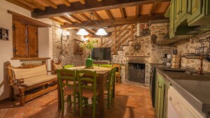 A rustic kitchen with a stone wall, wooden beams, a dining table with chairs, a television, and a fireplace.