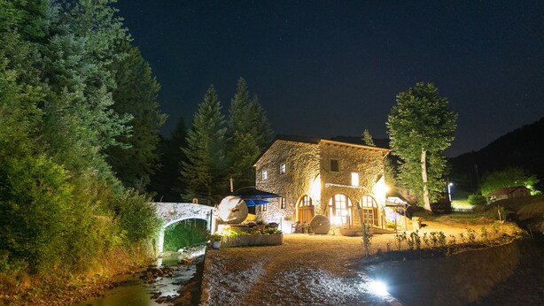 A stone house with a stone archway entrance, surrounded by trees and a small stream.