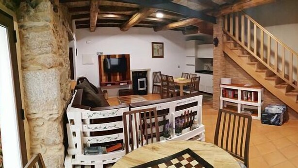 Un interior rústico con vigas de madera a la vista, una zona de comedor con mesa y sillas, una escalera y una pared de piedra.