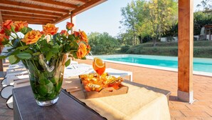 Ein Poolbereich mit einer Holzpergola, einer Glasvase mit orangefarbenen Rosen, einem Glas Orangensaft und einer Snackkörbchen.