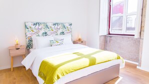 A neatly made bed with a floral headboard, two bedside tables with lamps, and a window with a view of buildings. - Spacious 3BR / 2 bath | Historic center Braga (Braga)