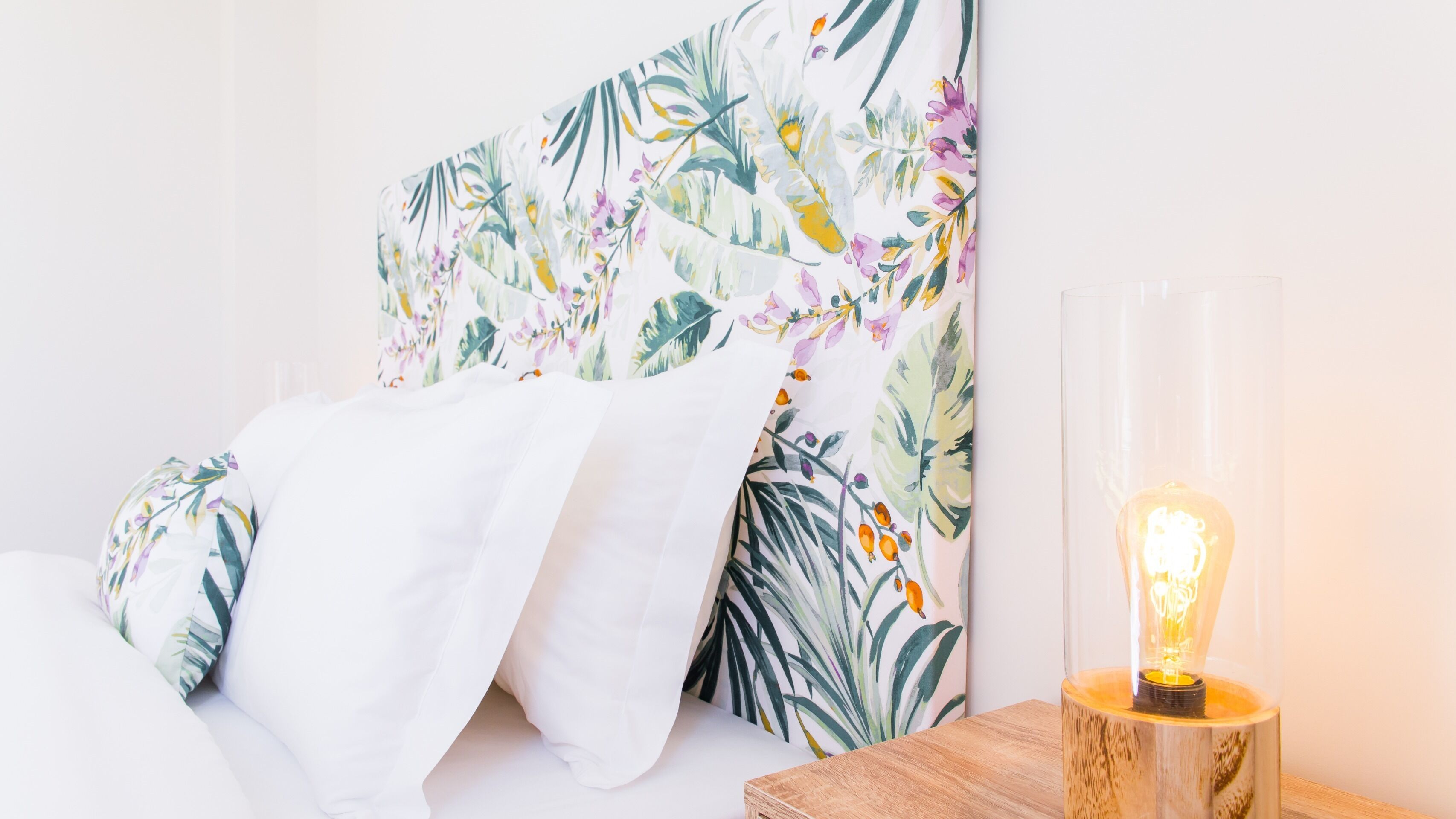 A bed with a floral-patterned headboard and a wooden bedside table with a lit bulb.