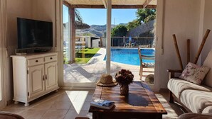 En stue med fjernsyn, spisebord med vase og magasin, sofa, samt udsigt til swimmingpool og udendørs område.