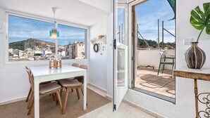 Een kamer met een eettafel en stoelen, een uitzicht op een stadslandschap door een groot raam, en een schuifdeur naar een buitenruimte met een stoel en een bloempot.