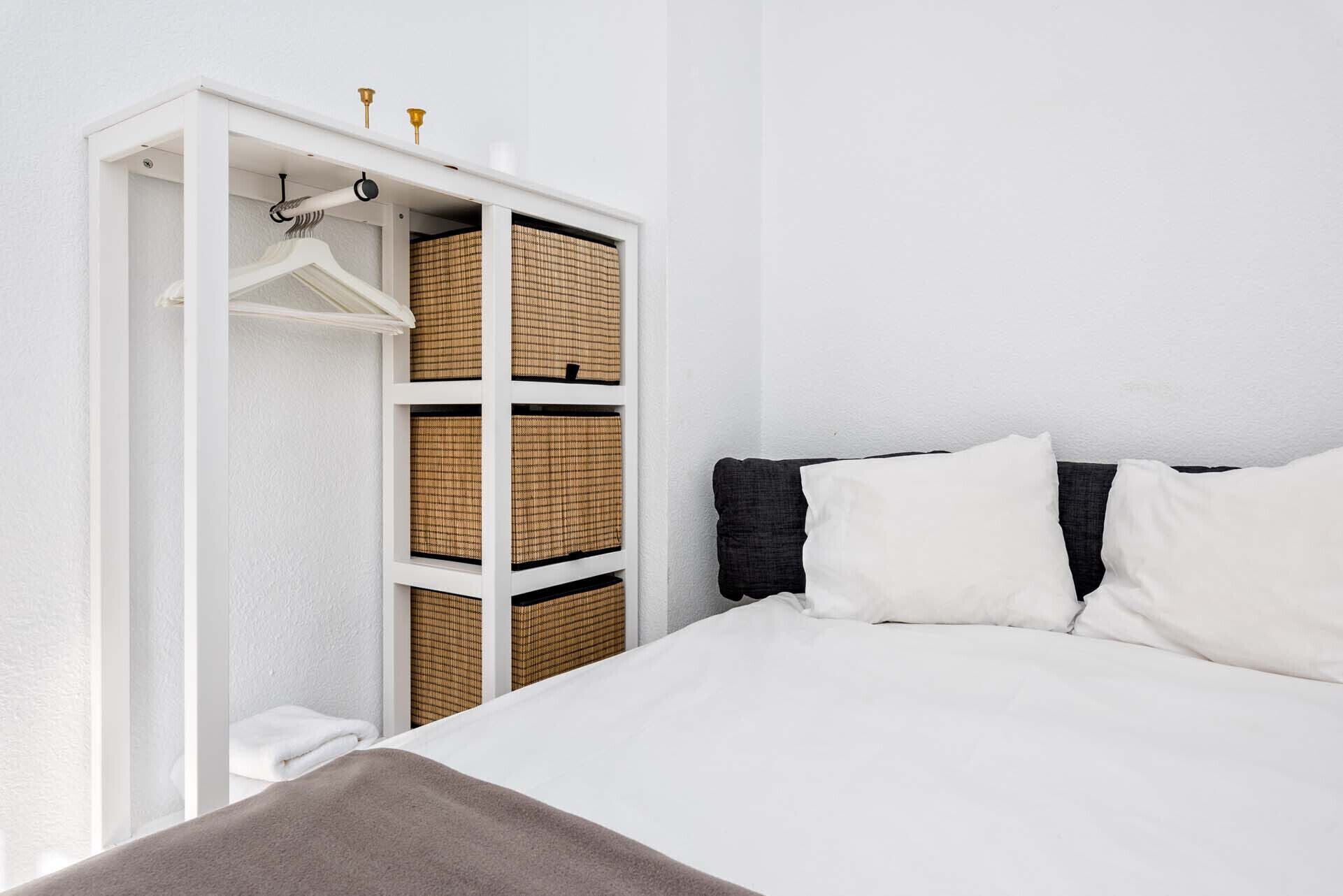 A bedroom with a bed, white bedding, a dark headboard, and a white wardrobe with baskets.