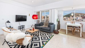 Een moderne woonkamer met een grijze bank, een houten salontafel, een televisie en een stadsgezicht door grote ramen.