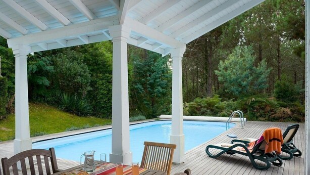 A covered outdoor area with a pool, wooden deck, lounge chairs, and a table with fruits and drinks.