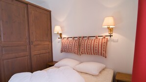 A bedroom with a bed, two bedside lamps, a headboard with patterned curtains, and a wooden wardrobe.