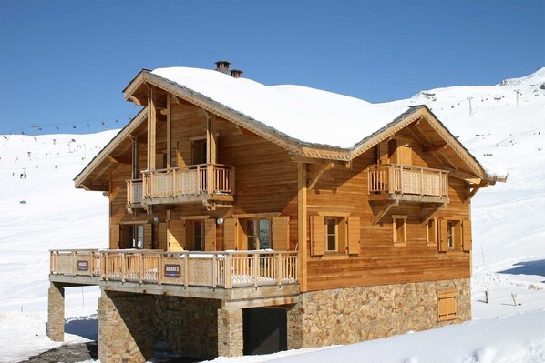 Un chalet en bois doté de balcons, avec une fondation en pierre et une enseigne indiquant « ARIANE II ».