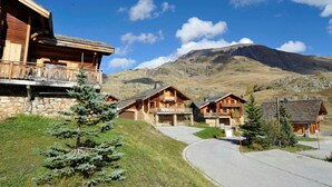 A mountain lodge with wooden cabins and a stone building, set against a backdrop of mountains and clear skies. - Chalet avec cheminée | Accès direct aux pistes ! (Huez)