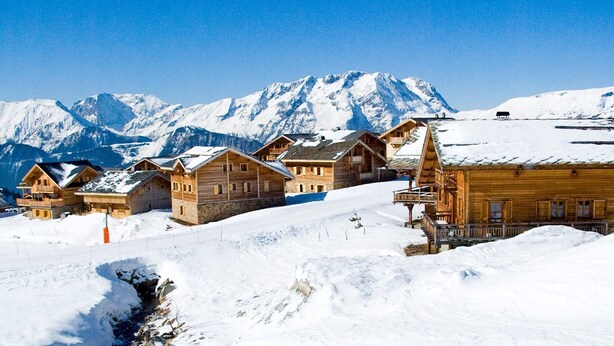 A row of wooden chalets with stone foundations, set against a backdrop of snow-covered mountains and a clear blue sky. - Chalet avec cheminée | Accès direct aux pistes ! (Huez)