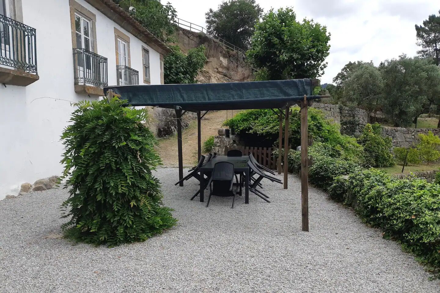 A patio with a table and chairs under a pergola, surrounded by gravel and greenery.