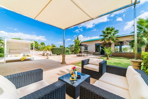 An outdoor patio with wicker furniture, a table with a drink, and a view of a house and palm trees. - Finca Son Perot with pool in Mallorca (Maria de la salut)