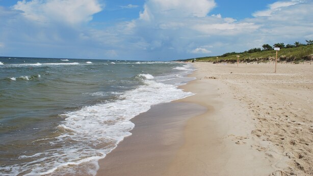 Ein sandiger Strand mit sanften Wellen, einem klaren Himmel und einem Schild, das den Zugang zum Strand weist.