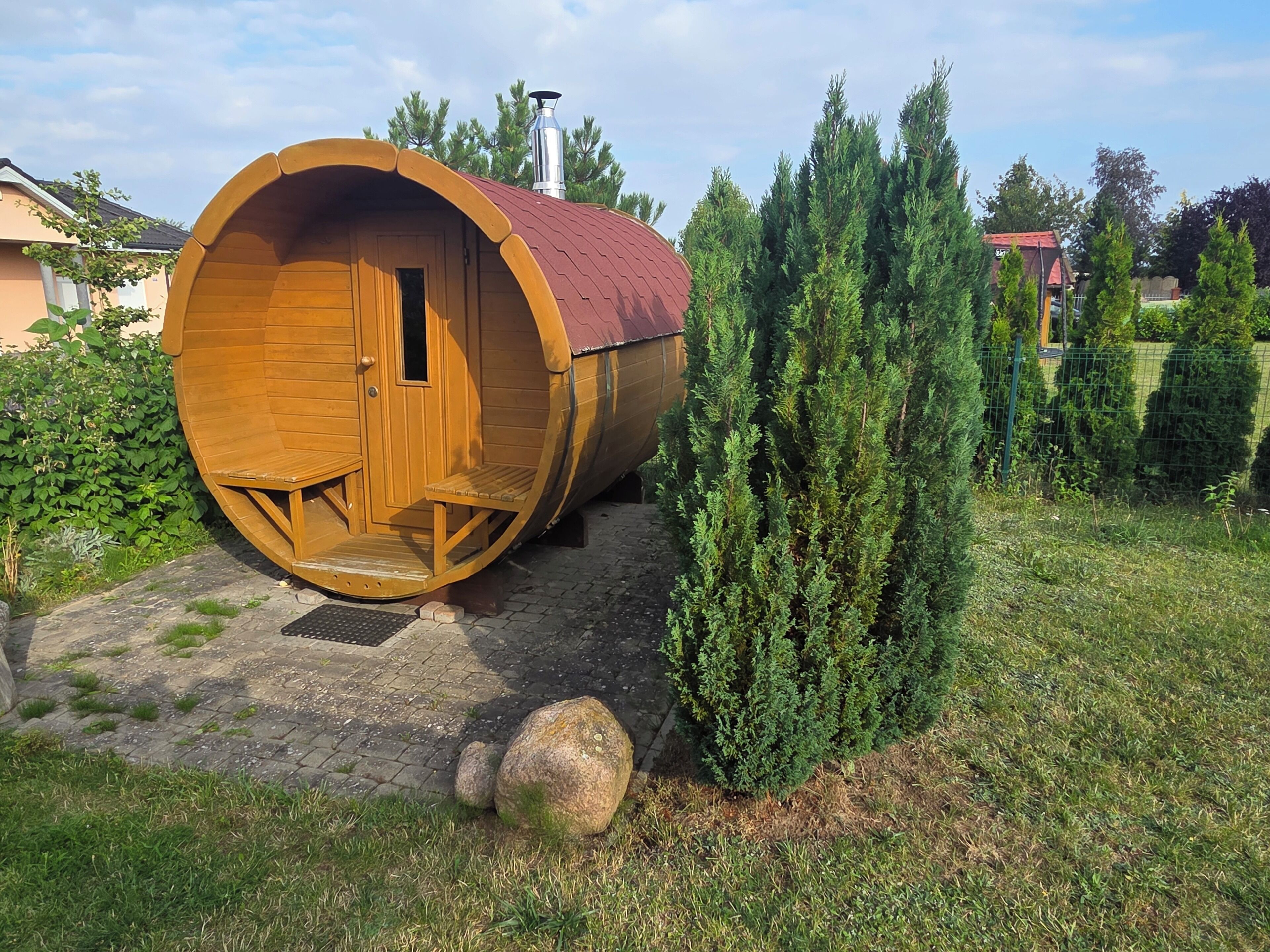 Un bâtiment en forme de tonneau, fait de bois, doté d’une porte et de fenêtres, entouré de verdure et bordé d’une zone pavée.