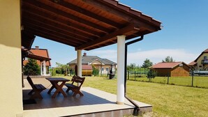 Eine überdachte Terrasse mit Tisch und Bänken, ein Rasen und Häuser im Hintergrund.