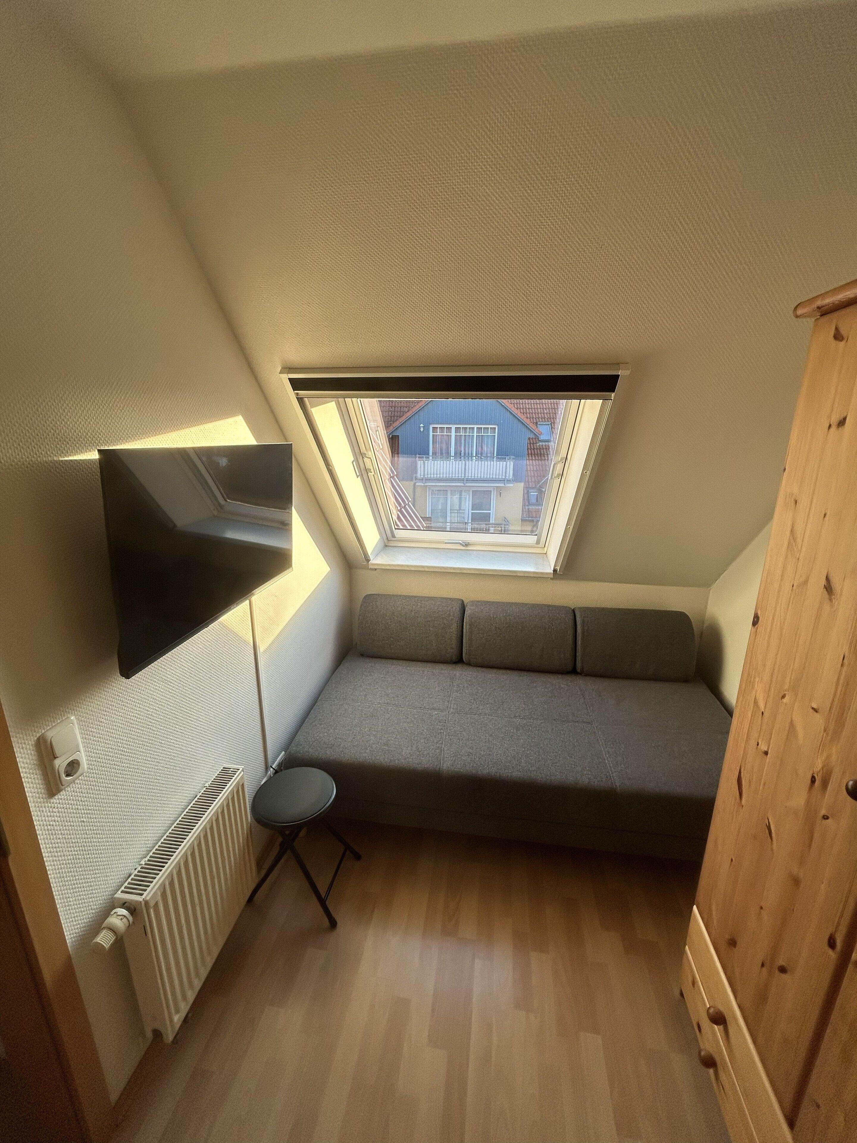 Ein kleiner Raum mit schrägem Dach, integriertem Fernseher, grauem Sofa, rundem Tisch, Heizkörper und Holzschrank.