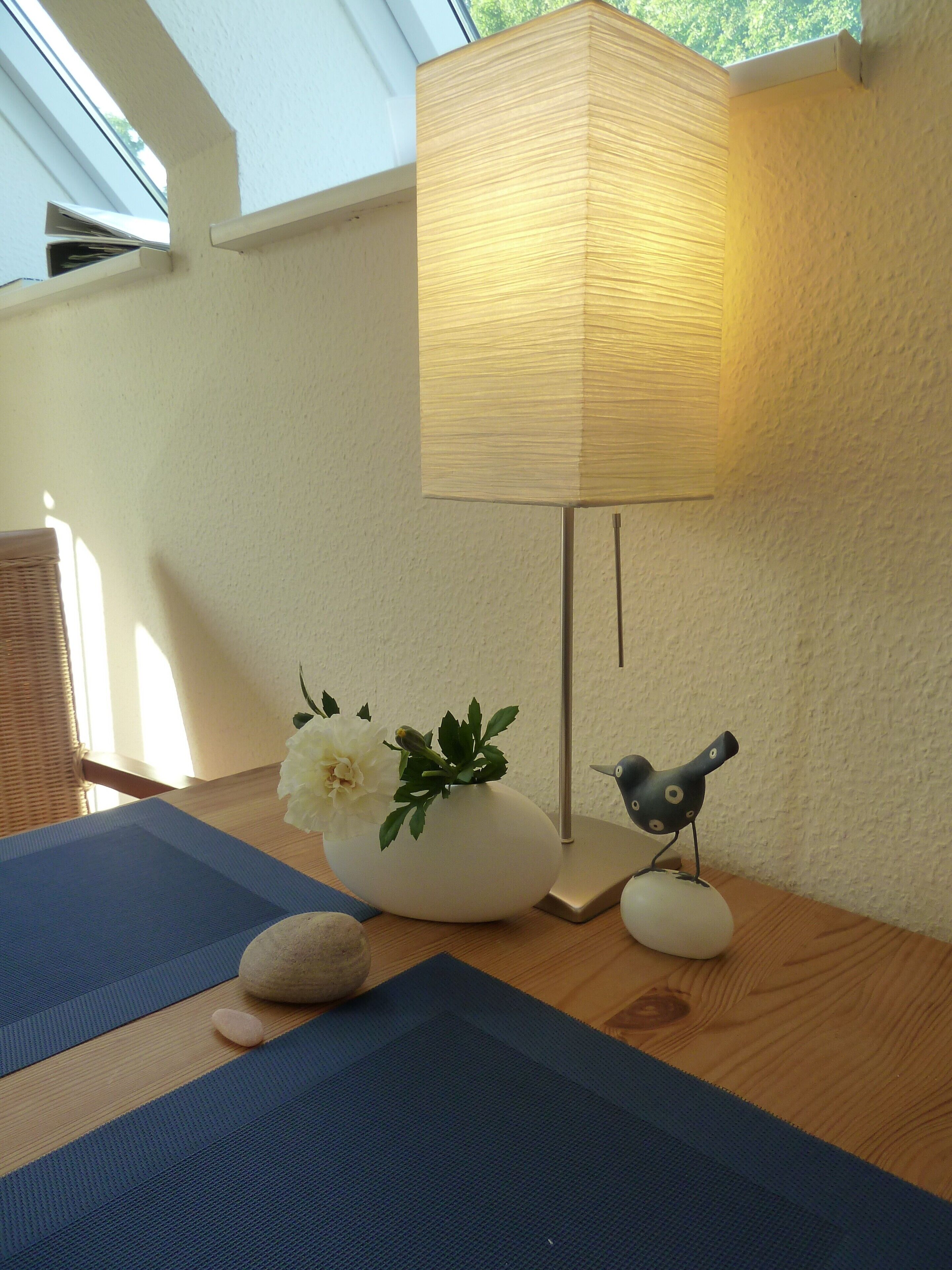 Una habitación con una mesa de madera, una lámpara, un jarrón con flores, una figurita de pájaro y un felpudo.