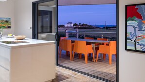 Une cuisine moderne dotée d’un grand îlot central, d’un coin repas avec des chaises orange et d’une vue sur la mer et les maisons.