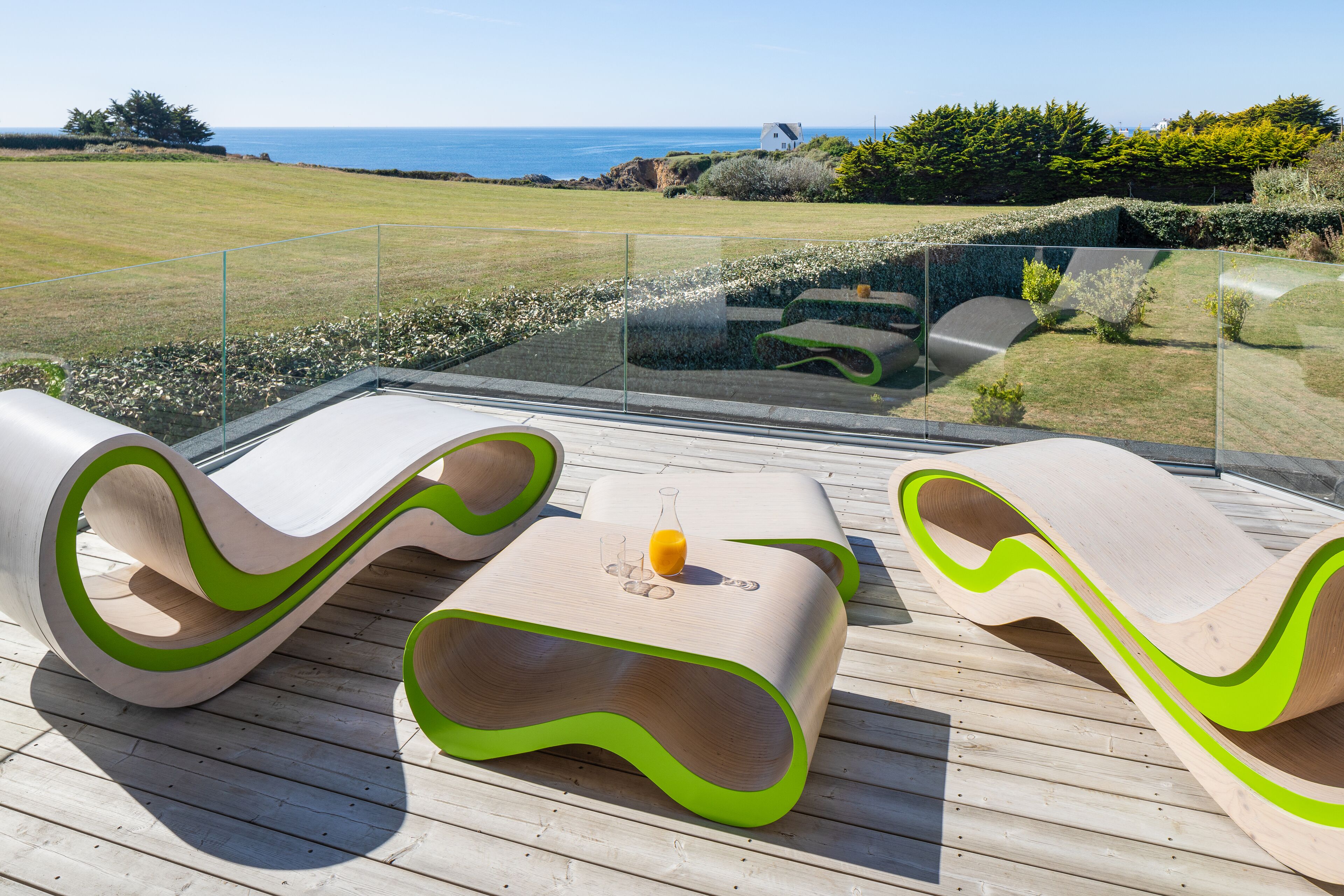 A deck with modern, wavy-designed lounge chairs and a glass table with a glass of orange juice.