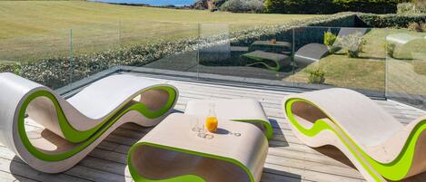 A deck with modern, wavy-designed lounge chairs and a glass table with a glass of orange juice.