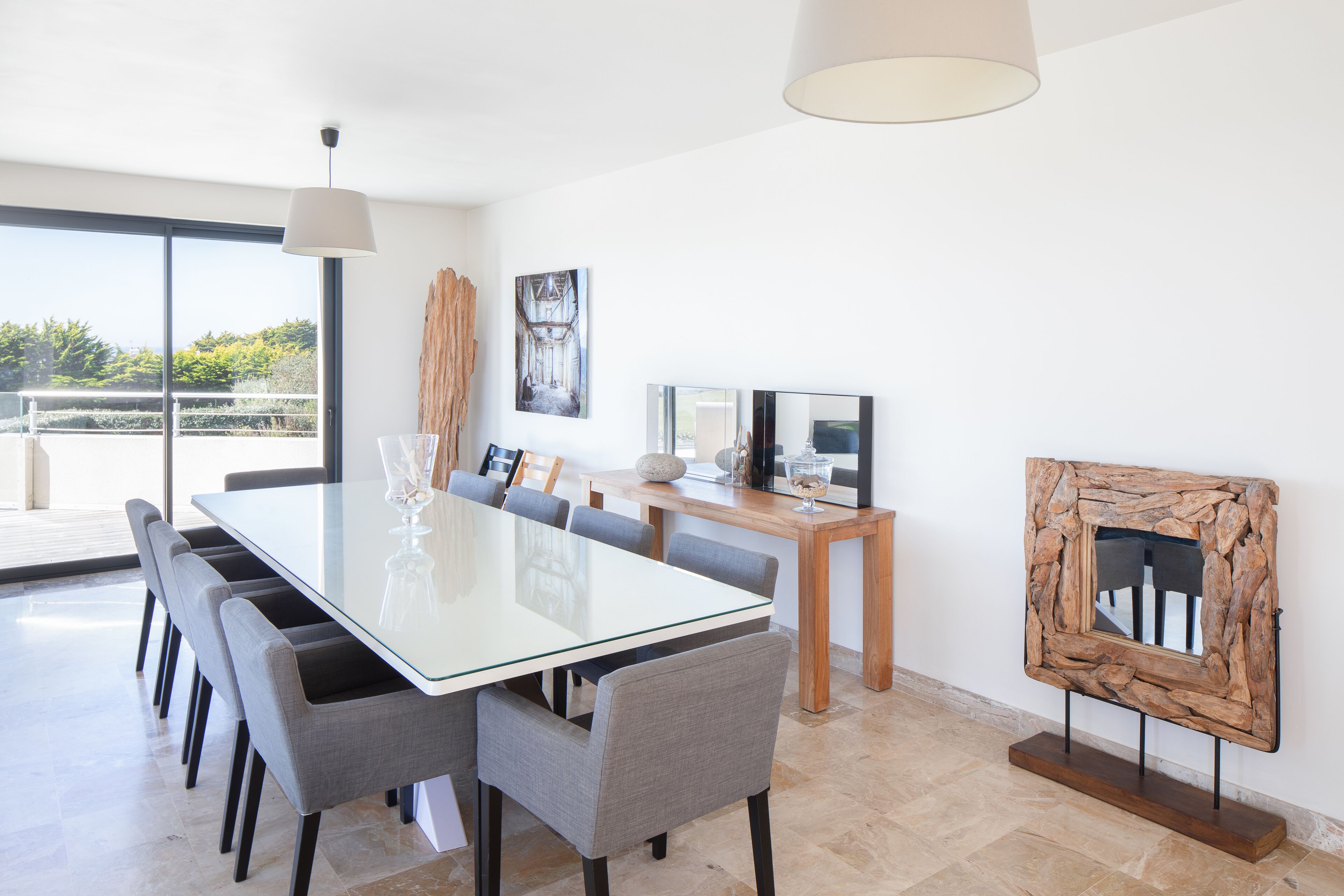 Un espace repas moderne avec une table en verre, des chaises grises et une table d’appoint en bois sur laquelle est posée une télévision.
