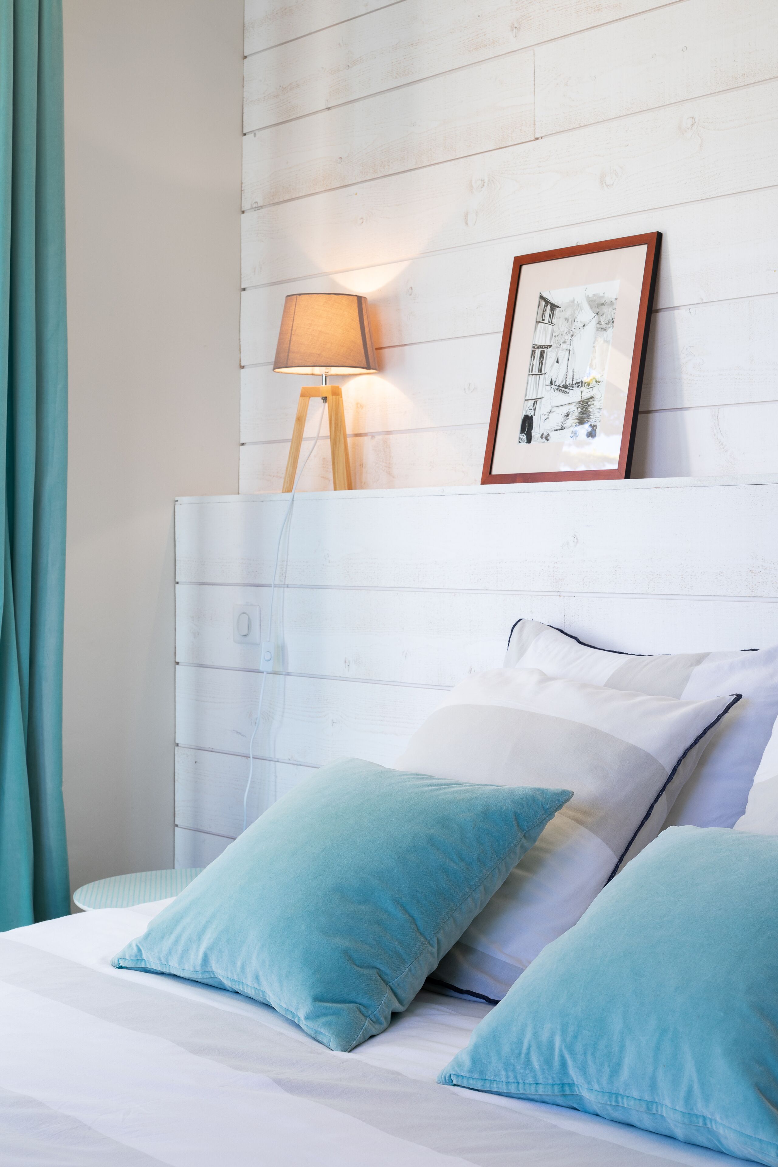 Una cama bien hecha con almohadillas azul claro, una lámpara de noche y un cuadro encolado sobre un cabecero de madera blanca.
