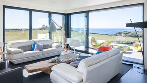 Un salon moderne avec un canapé blanc, un pouf bleu et une vue sur l’océan grâce à de grandes fenêtres.