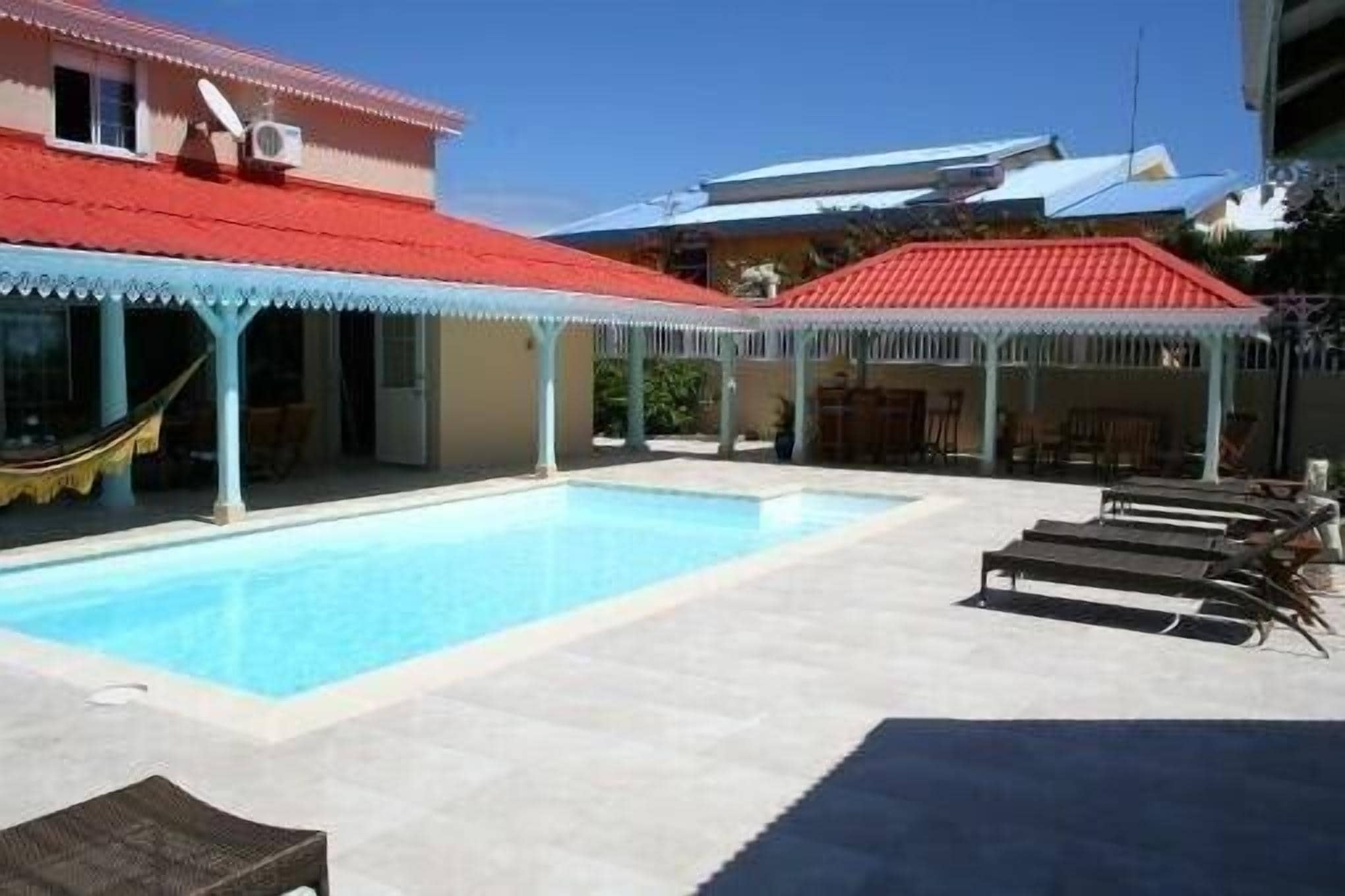 Una piscina con sillas de descanso, un área de estar techada y un edificio con techo rojo.