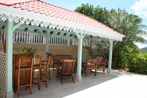 A covered patio area with wooden chairs and a table, surrounded by a white lattice fence and a red-tiled roof.