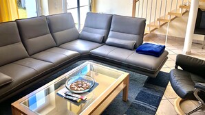 Una sala de estar con un sofá modular, una mesa de centro de vidrio con aperitivos, un sillón negro y una escalera.