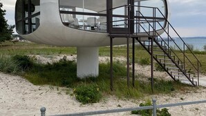 Una estructura moderna en forma de cúpula, con una escalera que conduce a un nivel superior, ubicada en una playa arenosa con vista al mar.