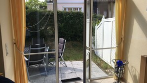 Una habitación con una puerta corrediza de vidrio que da a un patio con una mesa, sillas, un tapete azul y un soporte de metal con adornos.