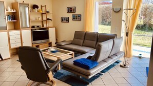 Una sala de estar con un sofá, una silla, un televisor, una estantería y una ventana con cortinas.