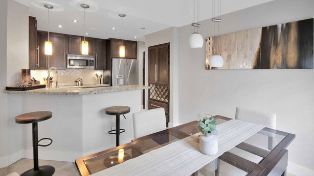 Una cocina moderna con armarios oscuros, un refrigerador de acero inoxidable y un microondas. Hay un comedor con una mesa de vidrio y dos taburetes de bar.