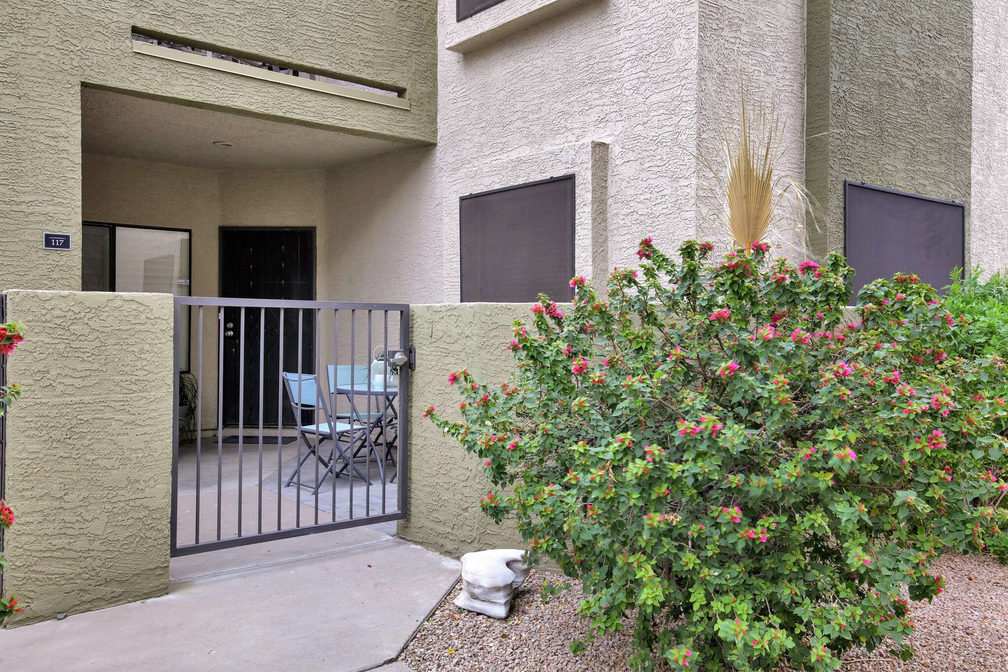 A building entrance with a gated patio, a small seating area, and a flowering bush.