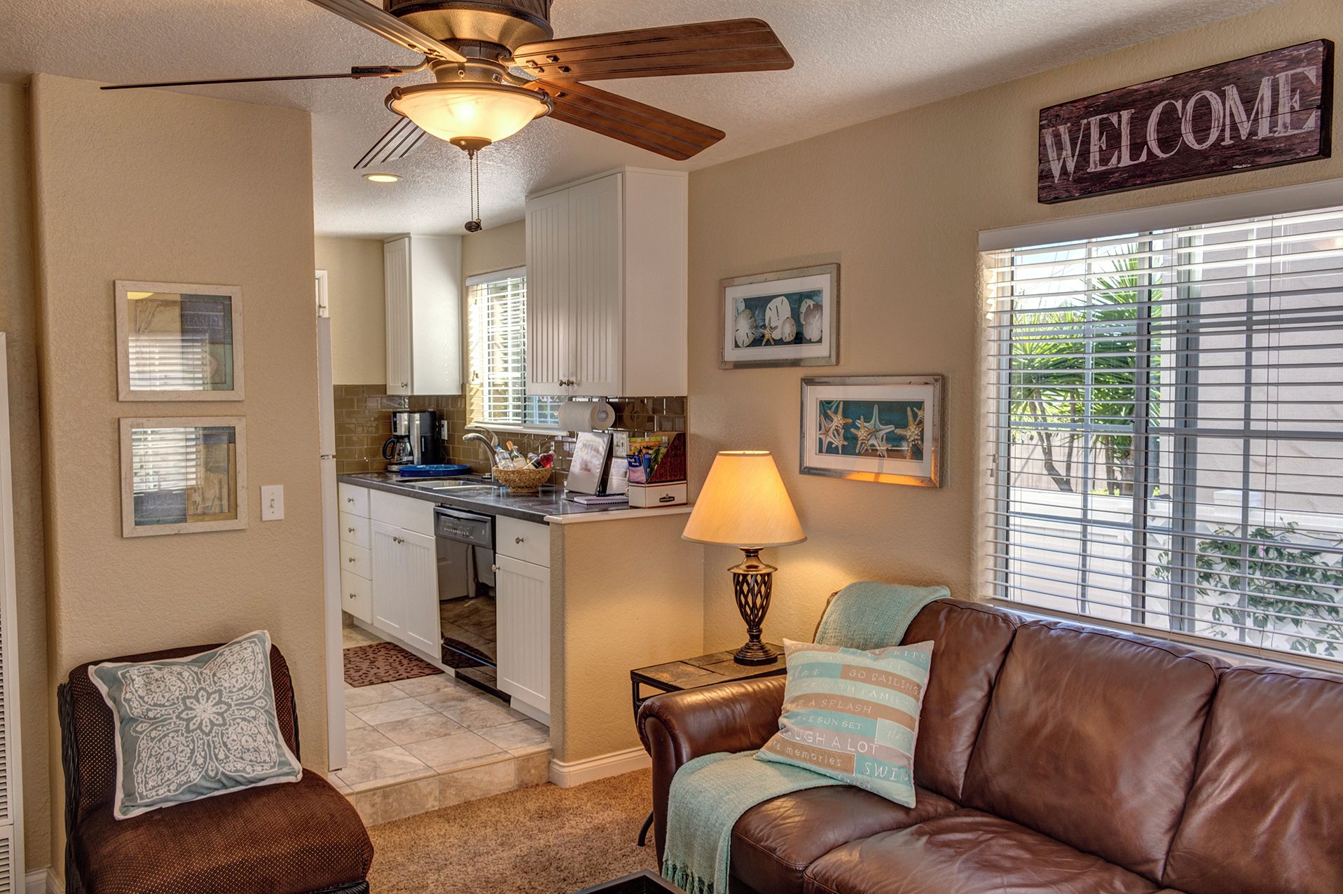 A compact living space with a kitchenette, a brown leather couch, a lamp, and a 'WELCOME' sign.