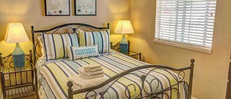 A neatly made bed with striped bedding and a pillow labeled "BEACH". There are two wall-mounted lamps on either side of the bed, and two framed artworks above the bed.