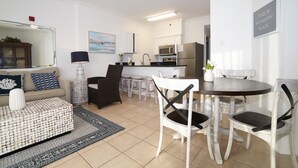 En moderne stue med sofa, sofabord og spiseområde med stole. Et køkken med hårde hvidevarer ses i baggrunden.