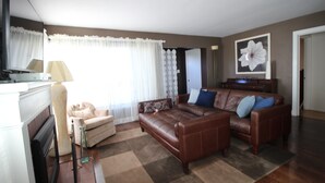Un salon avec un canapé en cuir marron, un fauteuil blanc, une cheminée et une grande fenêtre ornée de rideaux légers.
