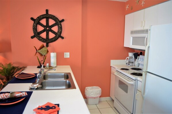 Una cocina con estufa, pileta y muebles blancos, una rueda decorativa y un bote de basura.