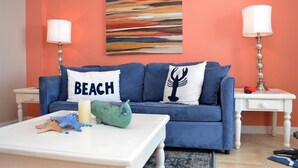 Ein blaues Sofa mit zwei Kissen, eines mit dem Aufdruck "BEACH" und das andere mit einem Baumdesign, ein weißer Couchtisch mit Spielzeug und zwei Lampen auf weißen Nachttischen vor einer orangefarbenen Wand.