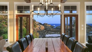 Uma área de refeições com mesa de madeira e cadeiras de couro, grandes janelas com vista para a costa rochosa e o oceano.