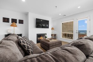 A modern living room with a fireplace, a flat-screen TV, and a balcony with a view of the sea.