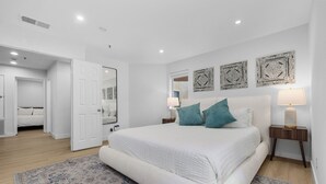 Une chambre spacieuse avec un grand lit, deux tables de chevet avec des lampes, un tapis décoratif et une porte donnant sur une autre pièce.