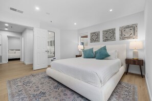 A spacious bedroom with a large bed, two bedside tables with lamps, a decorative rug, and a door leading to another room.