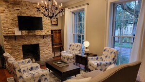A living room with a stone fireplace, two floral armchairs, a beige sofa, a coffee table, and a TV mounted on the wall. -  Jones St Hist Dist Gem 1853- 3KgBd/3Ba exquisite courtyard- 50 %offSun -ThurNts (Savannah)