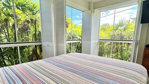 A bedroom with a large bed, a ceiling fan, and a view of lush greenery through the windows. - 
Kona coast studio, great rates free HS-WIFI, Parking, Walk to Magics Beach (Kailua-Kona)