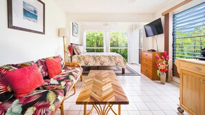 A bedroom with a bed, a sofa, a wooden table, a TV, and a view of greenery through the window. - 
Kona coast studio, great rates free HS-WIFI, Parking, Walk to Magics Beach (Kailua-Kona)